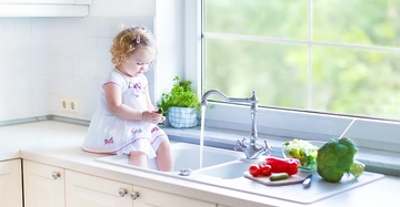 Ein kleines blondes Kind sitzt in einem weißen Kleid im Spülbecken, spielt mit Wasserhahn und grünem Kraut; umgeben von Küchenzeile mit Schneidebrett, Gemüse und Fensteraussicht ins Grüne.