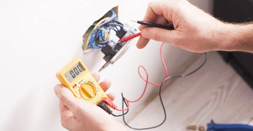 Ein Messgerät wird genutzt um elektrische Messungen an der offenen Steckdose in der Wand durchzuführen Spezielle Kabel sind im Gerät angeschlossen und die Umgebung ist ein Raum mit Holzboden