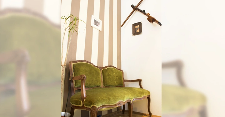 Galerie-Bild 5: Grünes antikes Sofa steht in einer Ecke mit gestreifter Tapete Eine Pflanze links und zwei Bilder sowie ein Gewehr an den Wänden
