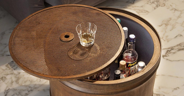 Ein runder Holztisch mit einem Gitterdeckel der angehoben ist. Ein Glas mit Getränk steht darauf. Innen sind mehrere Flaschen Alkohol zu sehen. Im Hintergrund ein Marmorboden und ein graues Sofa.