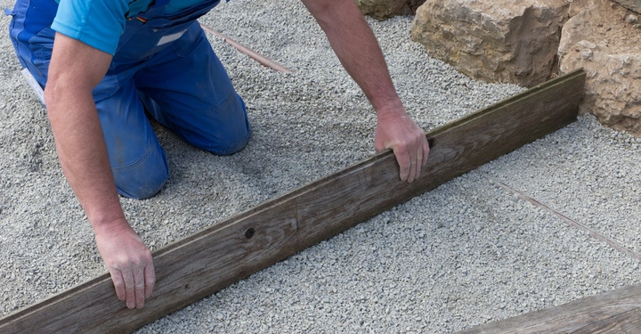 Eine Person in blauer Arbeitskleidung nivelliert eine Schicht Kies mit einem Holzbrett im Freien neben großen Felsen.