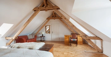 Bett und Sofa stehen in einem Dachzimmer mit sichtbaren Holzbalken hellen Holzfußboden und Schreibtisch mit Stuhl eine Lampe auf einem Beistelltisch neben dem Sofa.