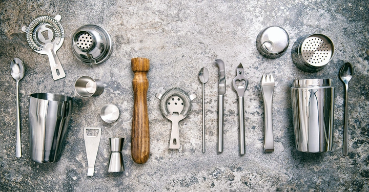 Verschiedene Barkeeper-Werkzeuge liegen ordentlich auf einer grauen, strukturierten Oberfläche. Die Werkzeuge sind aus Metall und Holz, darunter ein Cocktailshaker, ein Muddler und verschiedene Löffel und Siebe.