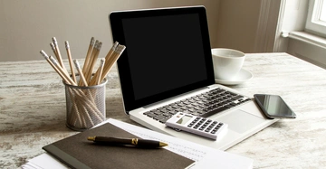 Laptop auf Schreibtisch steht neben einem Stiftebehälter Taschenrechner Notizbuch und Stift im hellen Raum mit Fenster im Hintergrund.