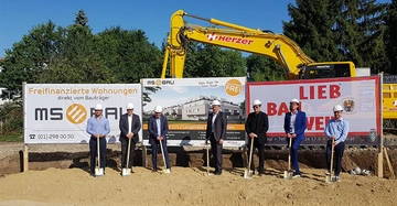 Eine Gruppe von sieben Menschen steht mit Schaufeln auf einer Baustelle; im Hintergrund sind drei Werbebanner und ein Bagger. Text: Freifinanzierte Wohnungen direkt vom Bauträger MS BAU Provisionsfrei. Telefonnummer (01) 298 00 50. www.msbau.cc Büro Wien. Lieb Bau Weiz.