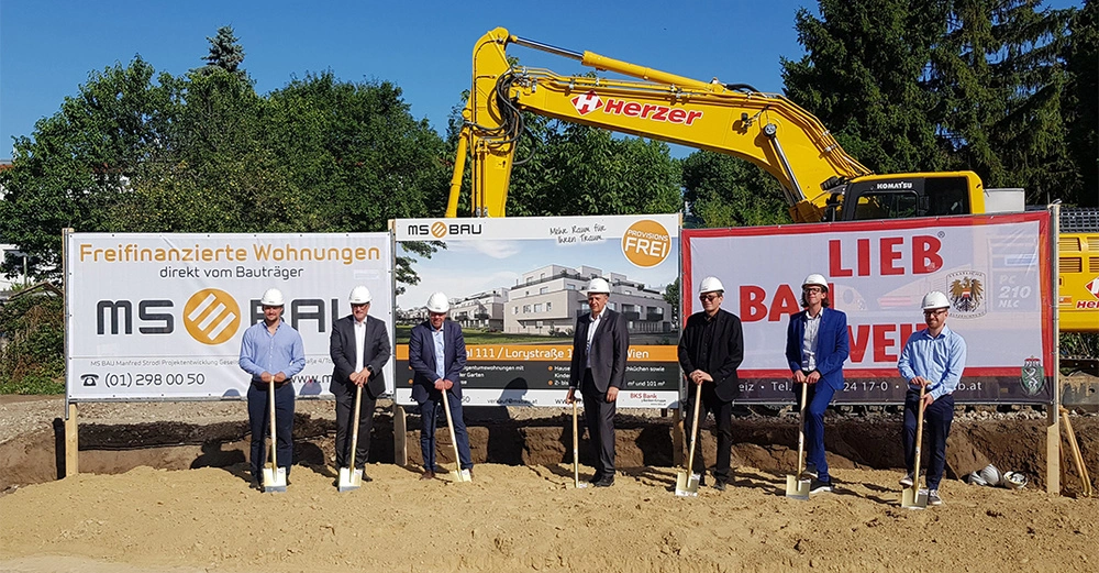 Eine Gruppe von sieben Menschen steht mit Schaufeln auf einer Baustelle; im Hintergrund sind drei Werbebanner und ein Bagger. Text: Freifinanzierte Wohnungen direkt vom Bauträger MS BAU Provisionsfrei. Telefonnummer (01) 298 00 50. www.msbau.cc Büro Wien. Lieb Bau Weiz.