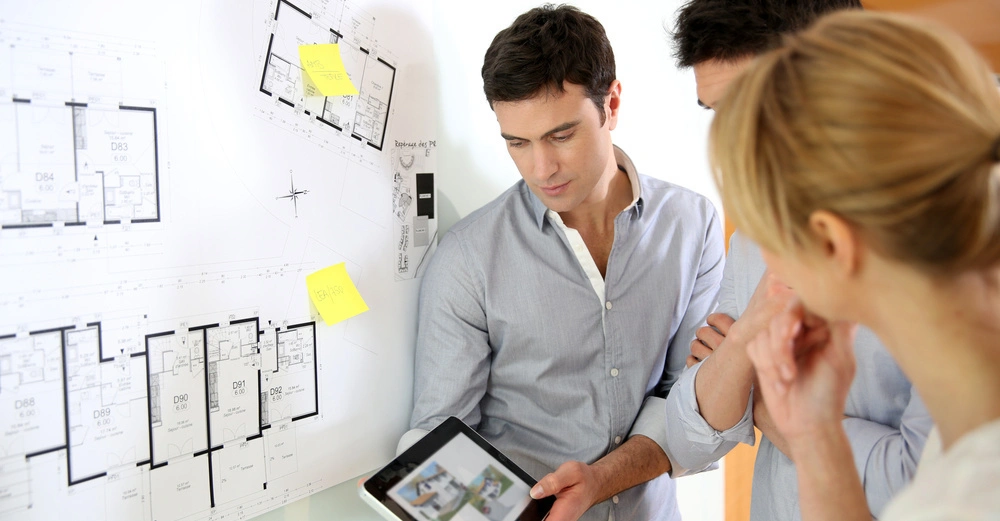 Drei Personen betrachten einen Tablet-Computer neben einem großen Architekturplan an der Wand eine Person zeigt auf das Tablet zwei gelbe Zettel stehen am Plan. Texte auf den Zetteln: Zettel 1 BEA + F05 Zettel 2 C+F + P.M.