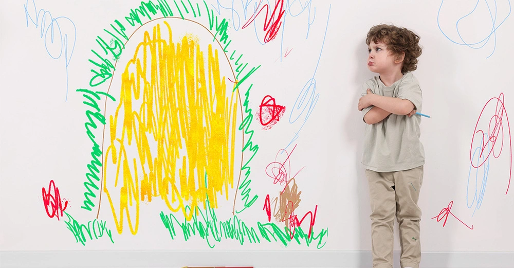 Verärgertes kleines Kind steht neben einer weißen Wand mit Kritzeleien. Flecken und Zeichnungen effektiv von Wänden entfernen.