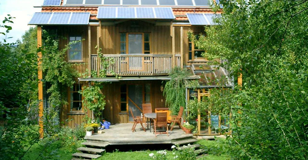 Ein zweistöckiges Holzhaus mit Solarpaneelen auf dem Dach steht in einem üppigen Garten. Eine Terrasse mit Tisch und Stühlen befindet sich im Vordergrund.