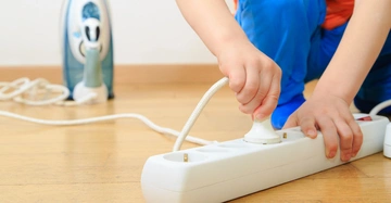 Kind zieht Stecker aus Mehrfachsteckdose auf Holzboden im Wohnbereich Bügeleisen und dessen Kabel im Hintergrund
