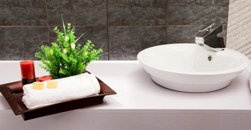 Weißes Waschbecken steht still auf einer weißen Badezimmerablage neben einem Dekotablett mit zwei Kerzen Handtuch und grünem Pflanzenarrangement im Badezimmer mit dunklen Fliesenwänden