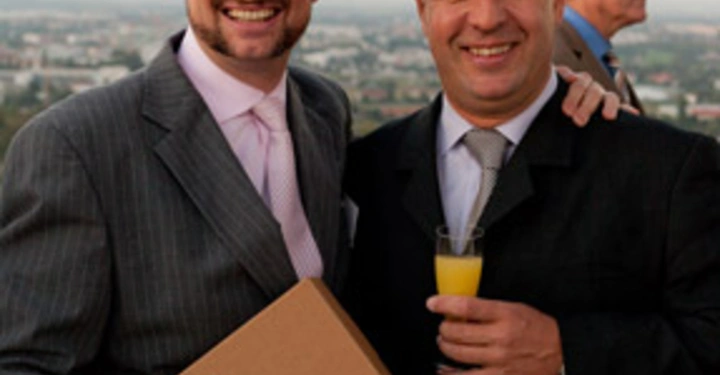 Galerie-Bild 45: Zwei Männer in Anzügen stehen nebeneinander und lächeln der eine hält eine Schachtel mit Weinflaschen der andere ein Glas Orangensaft ein Mann im Hintergrund betrachtet die Landschaft die Stadt im Hintergrund