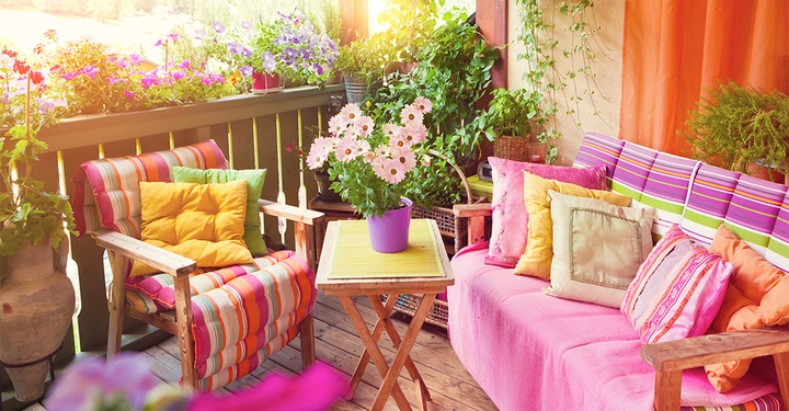 Blumenvase steht auf einem Holztisch Balkonmöbel sind bunt bezogen mit vielen Kissen Umgebung ist voller blühender Pflanzen in Töpfen und Hängekörben Sonnenlicht scheint durch die Pflanzen und dekoriert die Ecke