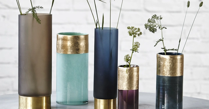 Galerie-Bild 2: Mehrere farbige Vasen mit Goldverzierung stehen auf einem Tisch, einige enthalten Zweige mit Blumen, in einem weißen Raum mit einer Ziegelwand im Hintergrund.