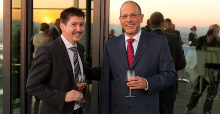 Galerie-Bild 35: Zwei Männer halten Gläser mit Getränken und lächeln auf einem Balkon bei Sonnenuntergang im Hintergrund weitere Menschen in formeller Kleidung stehend und plaudernd