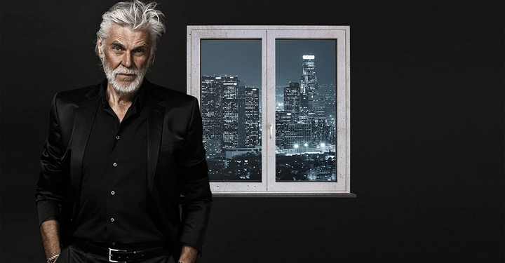 Mann mit grauem Haar und Bart steht in schwarzem Anzug neben einem Fenster mit Blick auf eine urbane Nachtszene mit beleuchteten Hochhäusern im Hintergrund.