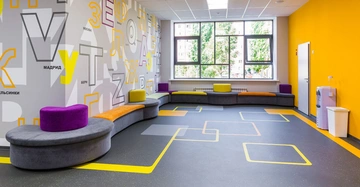 Bunte Sitzbänke stehen vor einer Wand mit großen Buchstaben und Wörtern wie MADRID MOSKAU und BERN in einem hellen Raum mit gelben Wänden und großen Fenstern