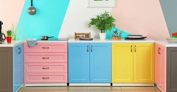 Küchenzeile mit drei Kästchen. Ganz links rosa, Mitte blau, rechts gelb. Die Wand dahinter ist in breiten Streifen in Türkis, Beige, Rosa und Grau gestrichen.