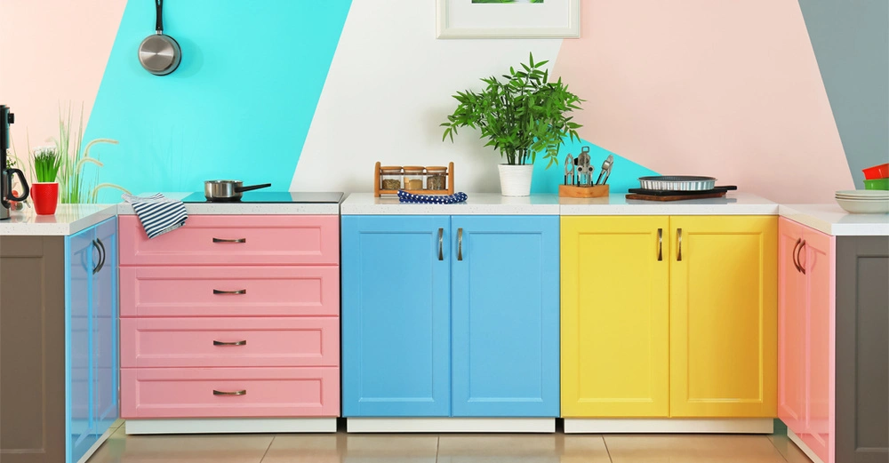 Küchenzeile mit drei Kästchen. Ganz links rosa, Mitte blau, rechts gelb. Die Wand dahinter ist in breiten Streifen in Türkis, Beige, Rosa und Grau gestrichen.