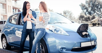 Zwei Frauen trinken Orangensaft und lehnen an einem blauen Elektroauto, das aufgeladen wird. Sie stehen auf einem städtischen Parkplatz an einem sonnigen Tag.