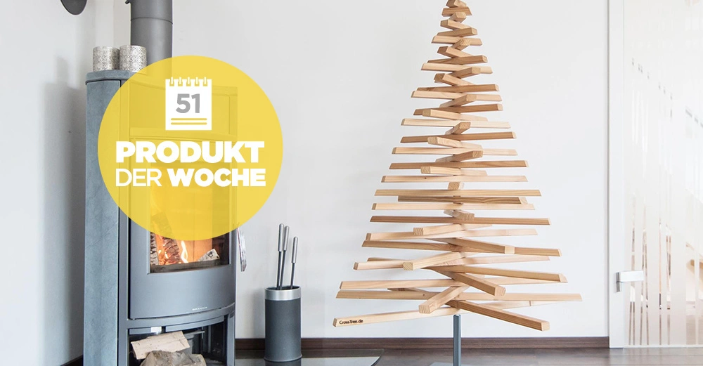 Ein moderner Holz-Weihnachtsbaum steht neben einem Kaminofen im Wohnraum. Text im Bild Produkt der Woche 51.