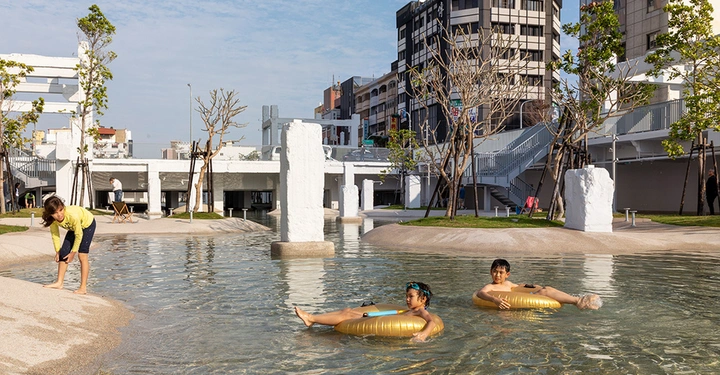 Galerie-Bild 6: Kinder spielen und schwimmen in einem flachen Pool in einer städtischen Parkanlage umgeben von Gebäuden Bäumen und Steinskulpturen in einer sonnigen Umgebung