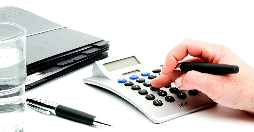 Hand drückt auf Taschenrechner in Büroumgebung mit Glas Wasser Aktenstapeln und Kugelschreiber