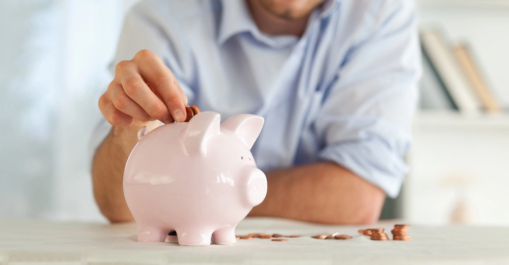 Rosa Sparschwein auf Tisch Mann steckt Münze hinein Im Hintergrund Bücherregal