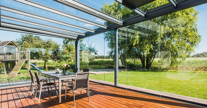 Galerie-Bild 5: Glastisch mit vier Stühlen steht auf einer überdachten Terrasse mit Glaswänden im Garten neben einem Spielhaus und Schaukeln umgeben von grünem Rasen und Bäumen bei sonnigem Wetter.