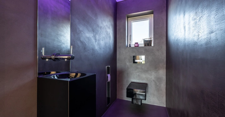 Galerie-Bild 7: Schwarzes Waschbecken steht an einer Wand mit modernem Wasserhahn. Raum mit dunkelgrauen Wänden und violetter Beleuchtung. Ein kleines Fenster mit Dekorationen oberhalb einer schwarzen Toilette. Text: LAMURISTA