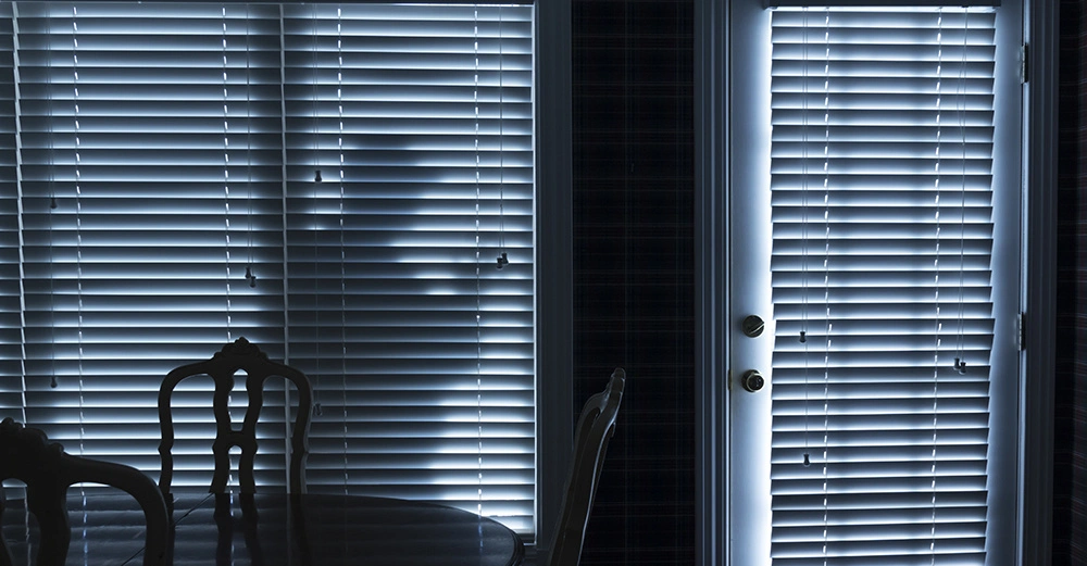 Holztisch und Stühle stehen im Vordergrund vor einer von Lamellenjalousien bedeckten Glastür Eine schemenhafte Figur erscheint hinter der Jalousie im Inneren eines dunklen Raums