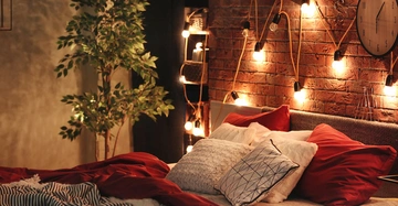 Bett mit roten und weißen Kissen ruht auf rot-weißer Bettdecke in gemütlichem Schlafzimmer mit hängenden Lichterketten an Ziegelwand und großer Pflanze daneben