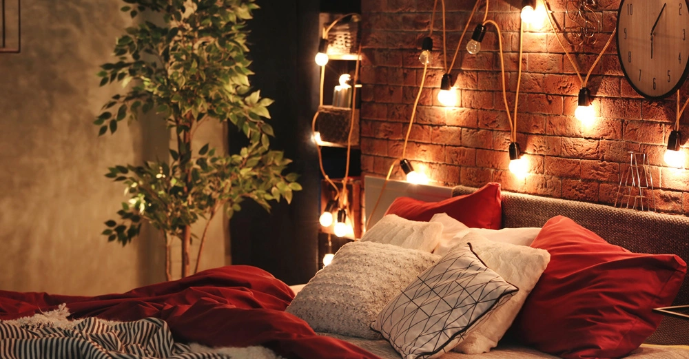 Bett mit roten und weißen Kissen ruht auf rot-weißer Bettdecke in gemütlichem Schlafzimmer mit hängenden Lichterketten an Ziegelwand und großer Pflanze daneben