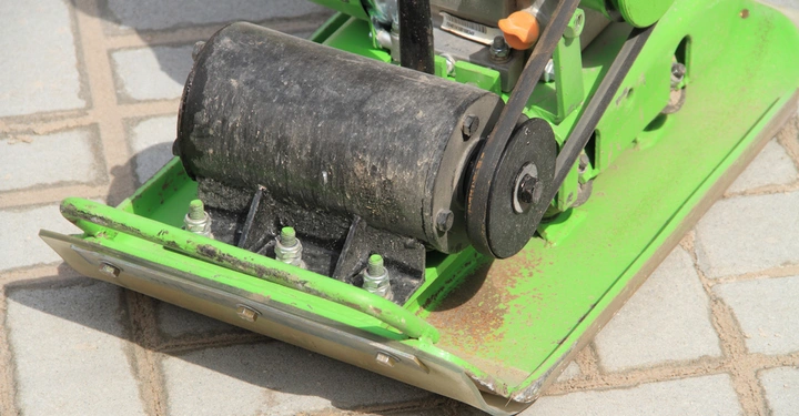 Grüne Rüttelplatte steht auf gepflastertem Boden. Staub und Schmutz sind um die Maschine herum sichtbar. Metallteile und Schrauben der Maschine heben sich durch ihre Funktion hervor.