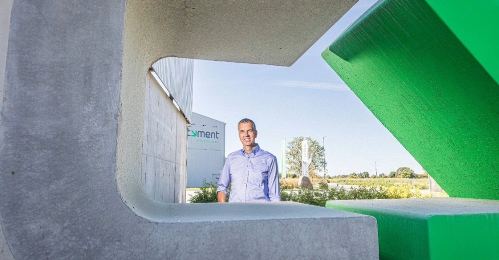 Porträt eines Mannes in einem hellblauen Hemd, der durch eine Öffnung aus massiven grauen und grünen Betonelementen blickt. Im Hintergrund ist ein Industriegebäude mit dem Logo "Cyment – building circular together" unter blauem Himmel zu sehen.