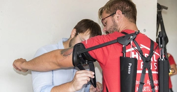 Ein Mann trägt ein schwarzes Exoskelett von Paexo während ein anderer Mann es justiert. Sie befinden sich in einem Innenraum. Text auf dem Hemd des Mannes: Hartl Arms Paexo Augsburg Hartl Am.