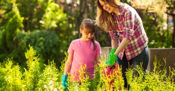 Frau gießt mit einem Mädchen in rosa T-Shirt Pflanzen in einem Garten. Die Umgebung ist grün und dicht bewachsen.