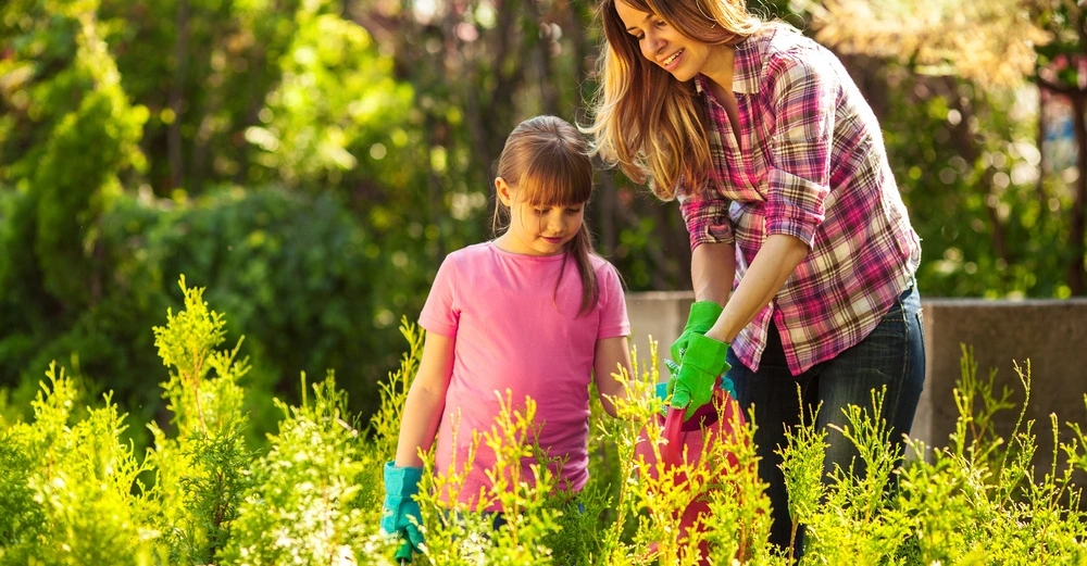 Frau gießt mit einem Mädchen in rosa T-Shirt Pflanzen in einem Garten. Die Umgebung ist grün und dicht bewachsen.