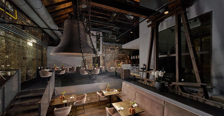 Große Glocke hängt von der Decke über Tischen in einem modernen Restaurant mit Backsteinwänden große Fenster Pflanzen und industriellen Maschinen im Hintergrund.