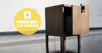 Holzschrank steht leicht geöffnet auf einem glänzenden Boden in einem modernen, minimalistischen Raum. Ein gelber Kreis enthält einen Kalender mit der Zahl 12 und den Text PRODUKT DER WOCHE.