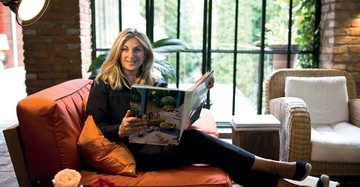 Frau liest ein Magazin auf einem roten Sofa in einem gut beleuchteten Raum neben einem großen Fenster und in der Nähe einer Zimmerpflanze und eines Korbsessels.