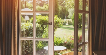 Offene Glastür führt zu einem sonnigen Garten mit üppigem grünen Rasen, Büschen und einem runden Tisch davor, umgeben von Vorhängen im Innenraum.