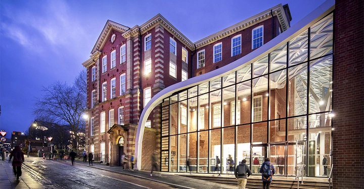 Galerie-Bild 1: Ein historisches Gebäude mit rotem Backstein und großen Fenstern steht an einer belebten, nassen Straße bei Dämmerung Menschen gehen vorbei große Glasfassade modernisiert einen Teil des Gebäudes Bäume im Hintergrund