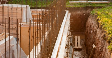 Baugerüst mit Stahlverstrebungen ragt aus einer Baugrube heraus. Im Hintergrund sind Zäune und Baumaterialien sichtbar, während im Vordergrund Erde und Gras den Rand der Grube säumen.