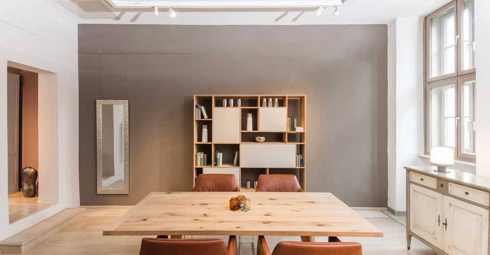 Holztisch steht in einem modernen Esszimmer zwischen braunen Stühlen und einem Holzregal. Umgeben von grauen Wänden und einem weißen Raum mit Fenstern, Spiegel und Sideboard.