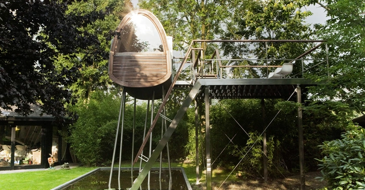 Galerie-Bild 2: Holzbau steht auf dünnen Stelzen über einer Wasserfläche in einem üppig grünen Garten mit hohen Bäumen und umgeben von Pflanzen. rechts befindet sich eine Treppe, die zur Plattform führt.