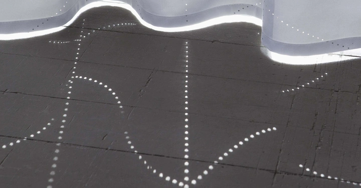 Galerie-Bild 10: Ein weißer Vorhang hängt und lässt punktuelle Lichtstrahlen durch kleine Löcher auf einen dunklen Holzboden fallen wodurch ein Muster erzeugt wird.