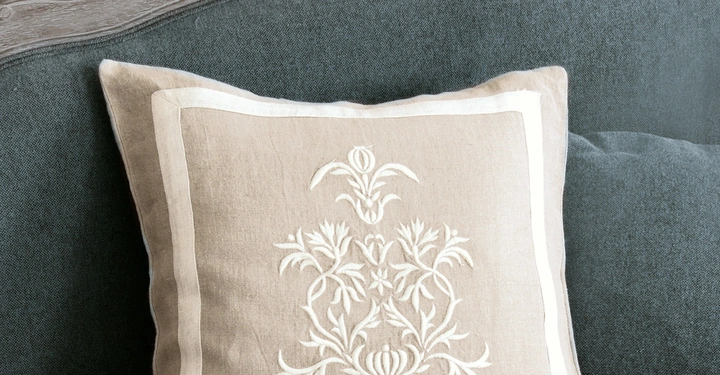 Galerie-Bild 4: Ein beiges Zierkissen mit weißem Stickmuster liegt auf einem grauen Sofa mit hölzerner Rückenlehne in einem Wohnzimmer.