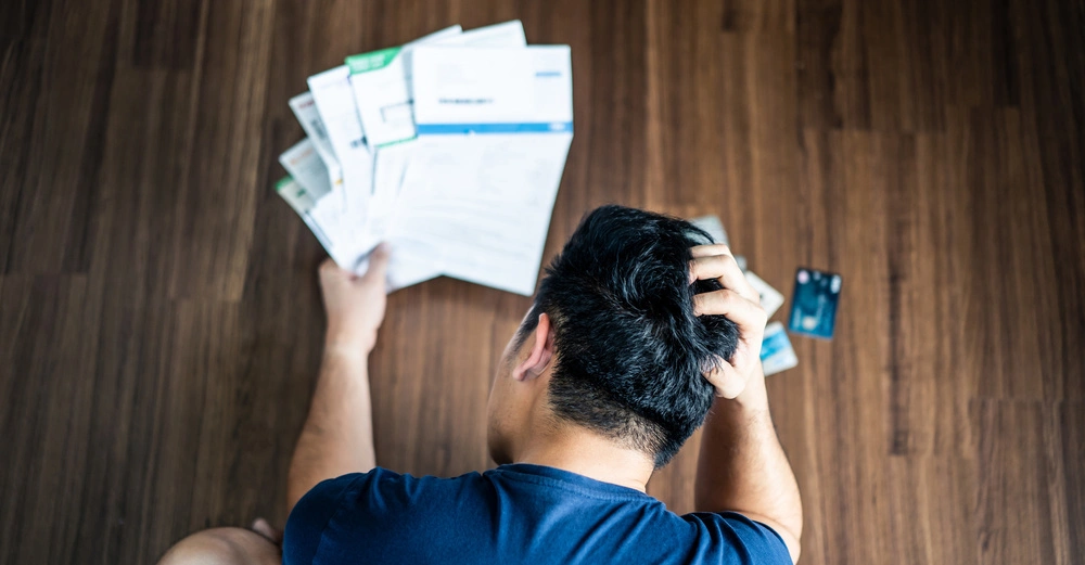Eine Person sitzt auf einem Holzboden hält Rechnungen in der Hand und kratzt sich am Kopf während Kreditkarten daneben liegen