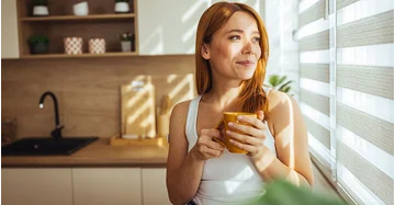 Frau vor einer Innenjalousie in einer gut beschatteten Küche mit modernem Sonnenschutz, hält eine Tasse und schaut lächelnd hinaus.